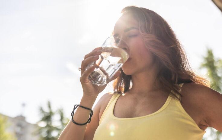 Frau in einem gelben Top genießt einen Schluck Wasser aus einem Glas im Sonnenlicht.