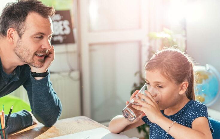 Ein kleines Mädchen trinkt mit beiden Händen ein Glas Wasser und ihr Vater schaut ihr dabei zu.