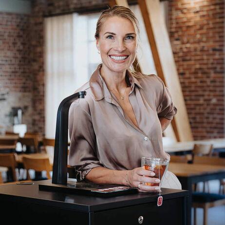 Eine Frau lächelt, während sie mit einem Teeglas in der Hand neben einem eleganten Wasserzapfhahn steht. Im Hintergrund sind Holzmöbel sichtbar.