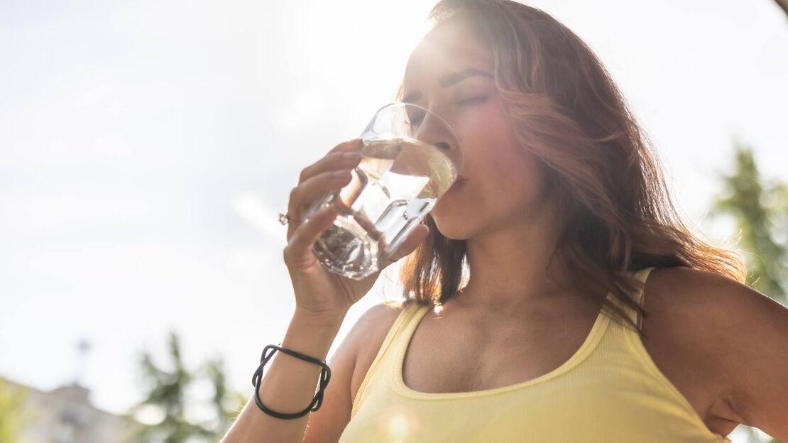 Frau in einem gelben Top genießt einen Schluck Wasser aus einem Glas im Sonnenlicht.