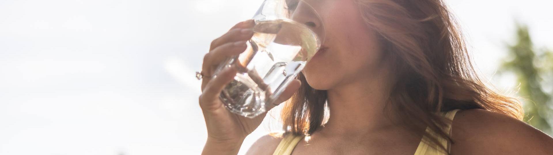 Frau in einem gelben Top genießt einen Schluck Wasser aus einem Glas im Sonnenlicht.