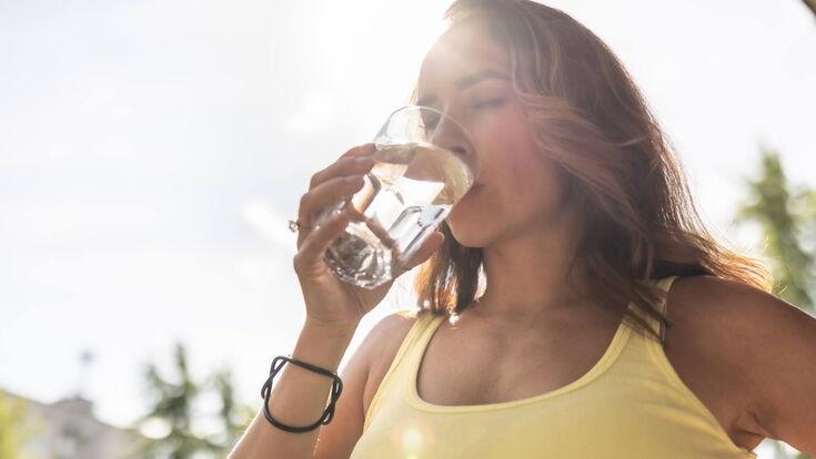 Frau in einem gelben Top genießt einen Schluck Wasser aus einem Glas im Sonnenlicht.