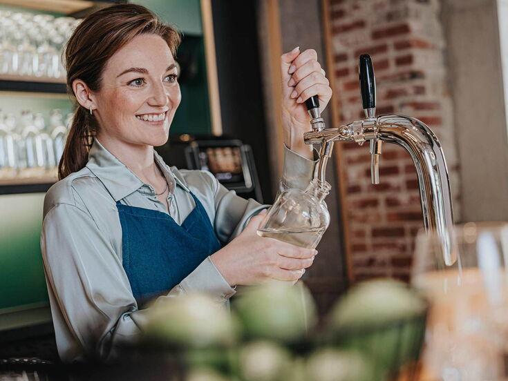 Eine freundliche Frau füllt frisches Wasser aus einem Wasserhahn in eine Glasflasche. Sie lächelt dabei. Der Hintergrund zeigt eine Bar- oder Restaurantumgebung.