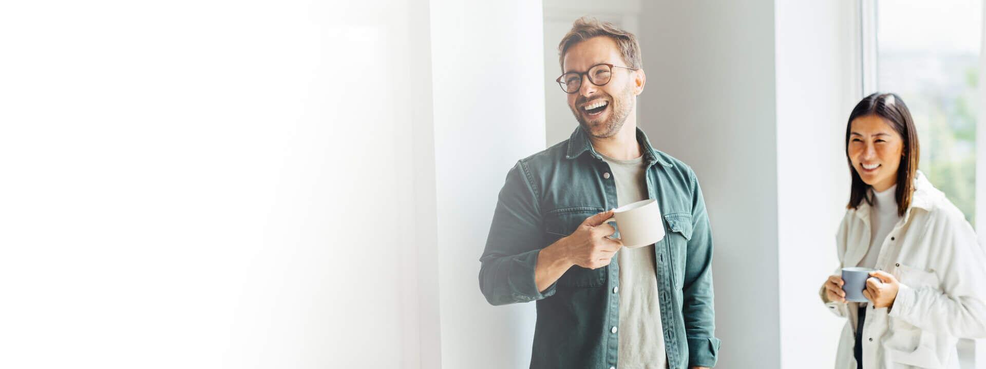 Zwei zufriedene Kunden stehen in einem hellen Raum und genießen eine Tasse Kaffee. Der Mann lacht und trägt eine Brille, während die Frau lächelnd ihre Tasse hält.