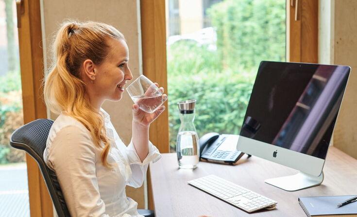 Frau sitzt am Schreibtisch mit einem Glas Wasser in der Hand