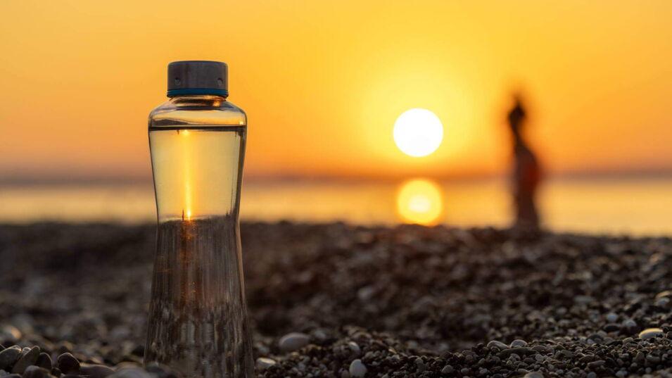 Eine durchsichtige Wasserflasche steht auf einem Kieselstrand, während die Sonne hinter ihr im Meer untergeht und ein goldener Lichtschein durch die Flasche hindurch scheint.