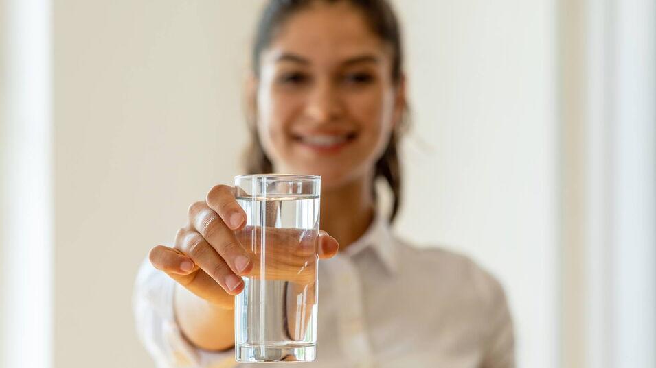 Junge Frau hält ein Glas Wasser in der Hand und lächelt in die Kamera.