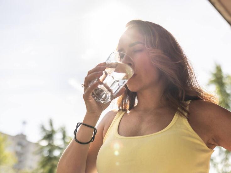 Frau in einem gelben Top genießt einen Schluck Wasser aus einem Glas im Sonnenlicht.